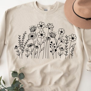 K&ouml;nnte beinhalten: Ein beiger Pullover mit einer schwarzen Linienzeichnung eines Blumenfeldes. Die Blumen sind stilisiert und beinhalten G&auml;nsebl&uuml;mchen, Wildblumen und andere Bl&uuml;ten.