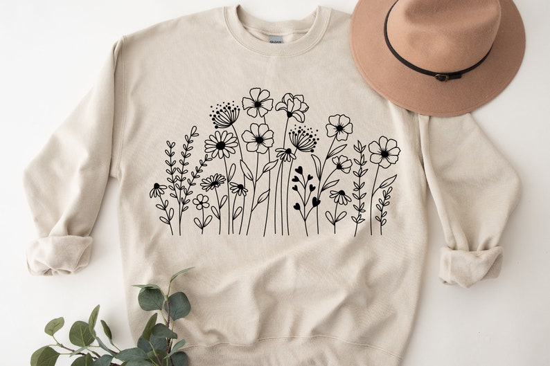 K&ouml;nnte beinhalten: Ein beiger Pullover mit einer schwarzen Linienzeichnung eines Blumenfeldes. Die Blumen sind stilisiert und beinhalten G&auml;nsebl&uuml;mchen, Wildblumen und andere Bl&uuml;ten.