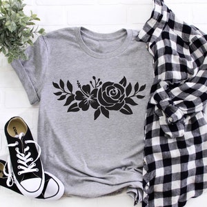 Pode incluir: Uma camiseta cinza com um design floral preto que apresenta uma rosa e um hibisco. A camiseta est&aacute; colocada sobre um fundo de tijolo branco com uma camisa xadrez branca e preta e t&ecirc;nis pretos.