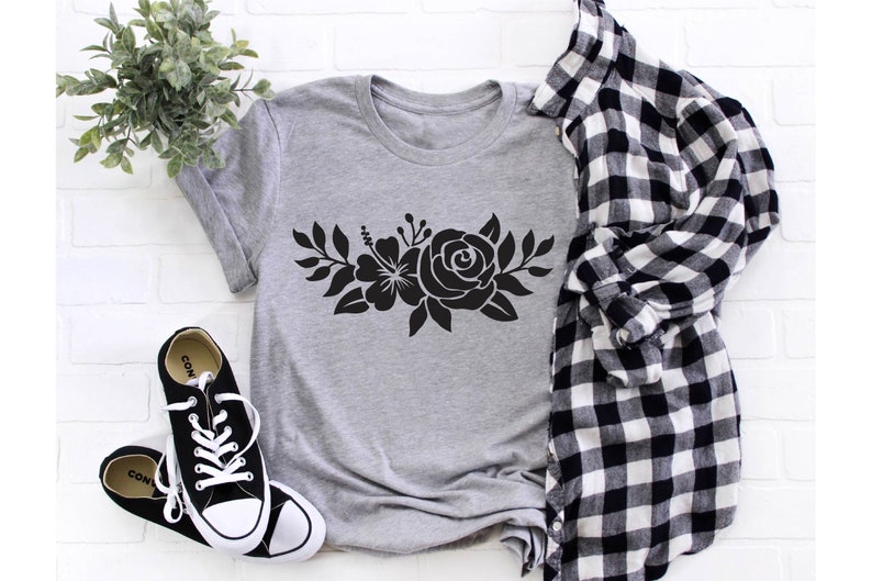 Pode incluir: Uma camiseta cinza com um design floral preto que apresenta uma rosa e um hibisco. A camiseta est&aacute; colocada sobre um fundo de tijolo branco com uma camisa xadrez branca e preta e t&ecirc;nis pretos.