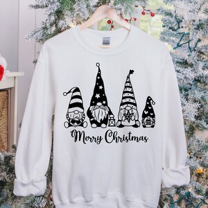 May include: White sweatshirt with a black graphic of four gnomes wearing striped hats and holding Christmas-themed items. The text "Merry Christmas" is printed below the gnomes.