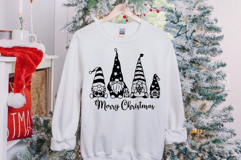 May include: White sweatshirt with a black graphic of four gnomes wearing striped hats and holding Christmas-themed items. The text "Merry Christmas" is printed below the gnomes.