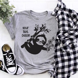 May include: Gray t-shirt with a black sloth design holding flowers and hanging from a branch. The text "Your Text Design" is printed on the shirt.