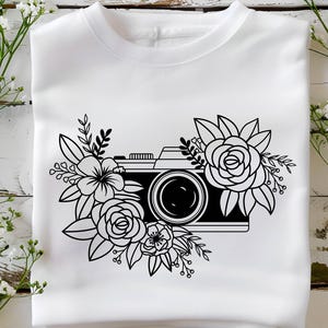 K&ouml;nnte beinhalten: Ein wei&szlig;es T-Shirt mit einer schwarz-wei&szlig;en Illustration einer Kamera, die von Blumen umgeben ist.
