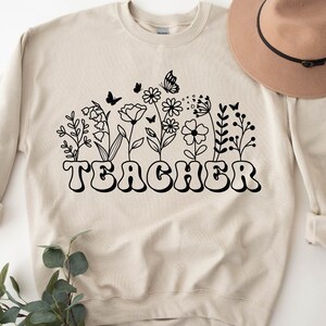 Może przedstawiać: Beżowa bluza z czarnym nadrukiem kwiat&oacute;w i motyli. Tekst "TEACHER" jest czarny i stylizowany czcionką retro.