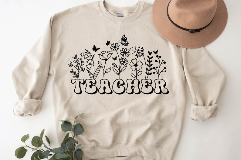 Może przedstawiać: Beżowa bluza z czarnym nadrukiem kwiat&oacute;w i motyli. Tekst "TEACHER" jest czarny i stylizowany czcionką retro.
