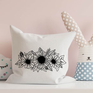 May include: A white cushion with a black floral design of sunflowers and leaves. The cushion is on a white shelf, with a stuffed bunny and cloud-shaped toy in the background.