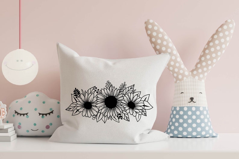 May include: A white cushion with a black floral design of sunflowers and leaves. The cushion is on a white shelf, with a stuffed bunny and cloud-shaped toy in the background.