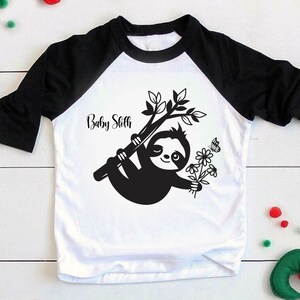 May include: White and black baseball tee shirt with a black sloth holding flowers and a butterfly. The text "Baby Sloth" is printed on the shirt.