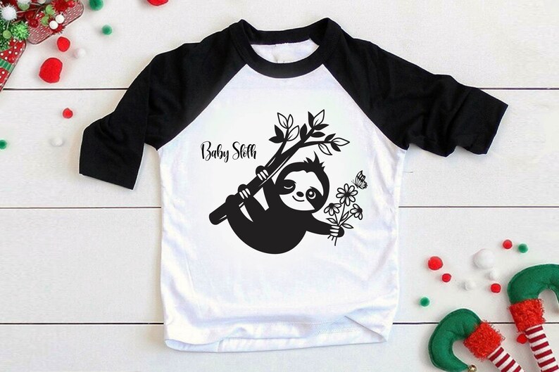 May include: White and black baseball tee shirt with a black sloth holding flowers and a butterfly. The text "Baby Sloth" is printed on the shirt.