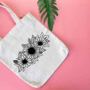 May include: Cream canvas tote bag with a black and white floral design of sunflowers and leaves. The tote bag has two white handles and is set against a pink background with a green leaf.