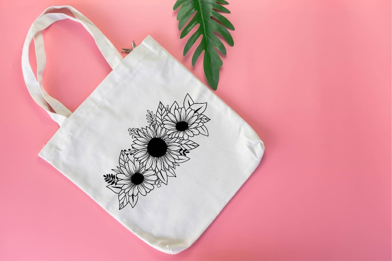 May include: Cream canvas tote bag with a black and white floral design of sunflowers and leaves. The tote bag has two white handles and is set against a pink background with a green leaf.