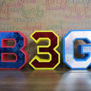 Puede incluir: Tres letras grandes y coloridas que deletrean "B3G" est&aacute;n hechas de cart&oacute;n y decoradas con purpurina y patrones. Las letras est&aacute;n dispuestas en una fila sobre una superficie de madera.