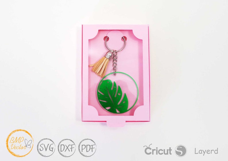 May include: A green monstera leaf keychain with a brown tassel charm in a pink box. The keychain is made of acrylic and has a silver metal ring.