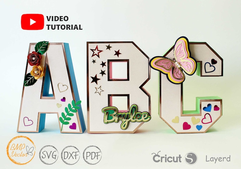 以下が含まれることがあります： 装飾的な装飾が施された3次元紙文字で「ABC」と綴られています。文字「A」には花と葉、文字「B」には星と「Brylee」という名前、文字「C」には蝶とハートがあります。