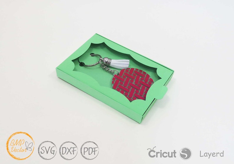 May include: A green box with a cut-out window, showcasing a red and white keychain with a tassel. The keychain is made of a woven material and has a silver metal ring.