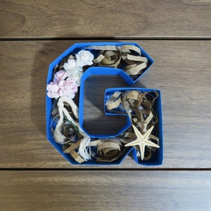 Puede incluir: Una letra G de cart&oacute;n azul llena de virutas de papel marr&oacute;n, flores blancas y rosas y una peque&ntilde;a estrella de mar.