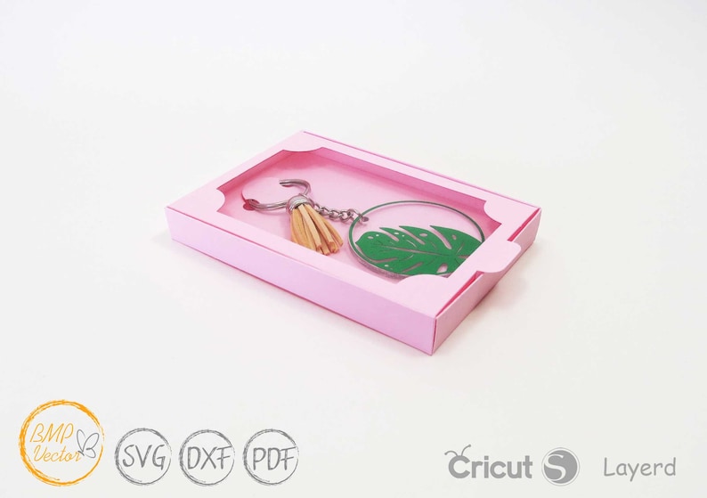 May include: A pink box with a clear window, showcasing a green leaf keychain with a tassel. The keychain is on a silver chain.