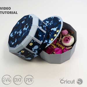 May include: A blue and white camouflage patterned paper craft box with a lid. The box is shaped like a hat and has a gold star design on the lid. The box is open and filled with pink and white tissue paper, pink candy, and pink flowers.