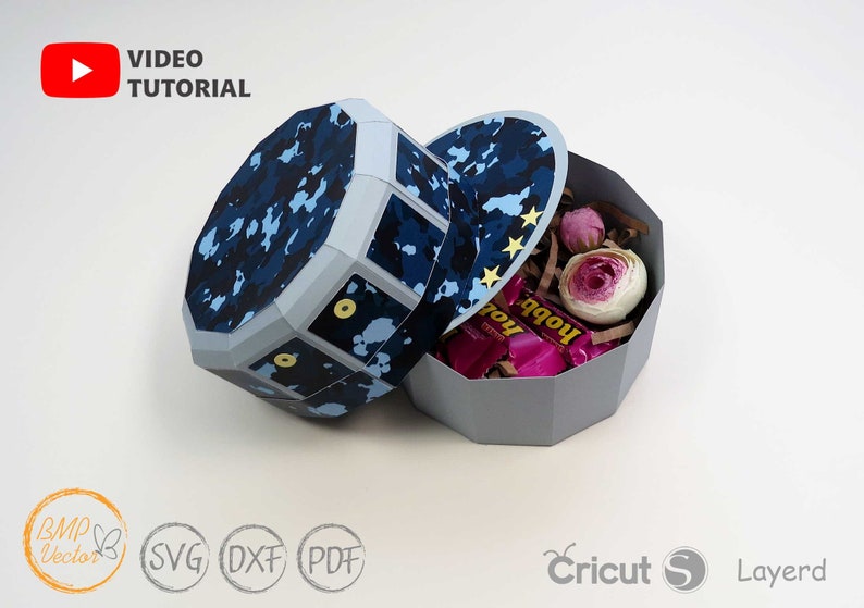 May include: A blue and white camouflage patterned paper craft box with a lid. The box is shaped like a hat and has a gold star design on the lid. The box is open and filled with pink and white tissue paper, pink candy, and pink flowers.