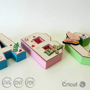 以下が含まれることがあります： 装飾的な要素で飾られた、3 次元の紙製の文字「ABC」。「A」は青で、花と葉で飾られています。「B」はピンクで、星と「Brylee」という名前で飾られています。「C」は緑で、蝶とハートで飾られています。