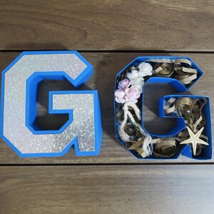 Puede incluir: Dos letras de cart&oacute;n azul, cada una con forma de la letra G. La letra de la izquierda est&aacute; cubierta de purpurina plateada. La letra de la derecha est&aacute; llena de art&iacute;culos decorativos, incluyendo flores, cuerda y una estrella de mar.