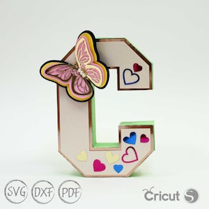 以下が含まれることがあります： ピンクの蝶とカラフルなハートが付いた、3Dレイヤーペーパークラフトの文字C。文字はベージュで、銅色のトリムと緑色の裏地が付いています。