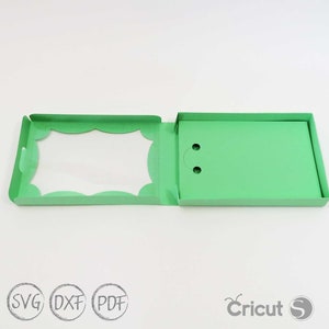 May include: A green card with a scalloped edge and a window for a photo. The card is open to reveal a green inner flap with two small holes.