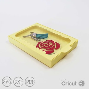 May include: A yellow cardboard box with a clear window, showcasing a red rose keychain with a teal tassel. The box has a scalloped edge and the text "Cricut S Layerd" is visible on the bottom right corner.
