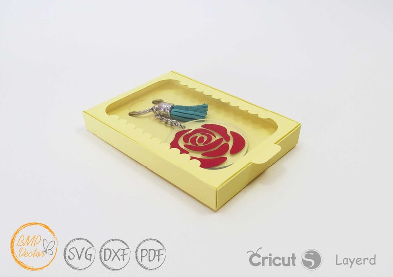 May include: A yellow cardboard box with a clear window, showcasing a red rose keychain with a teal tassel. The box has a scalloped edge and the text "Cricut S Layerd" is visible on the bottom right corner.