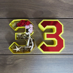 Puede incluir: Dos n&uacute;meros amarillos, 33, con un interior rojo. El n&uacute;mero 3 de la izquierda est&aacute; lleno de peque&ntilde;os objetos decorativos, incluyendo flores, cuentas y una estrella de mar.