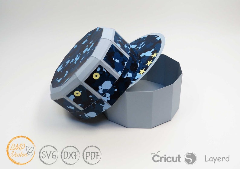 May include: A blue and white camouflage patterned paper box with a lid shaped like a baseball cap. The lid has yellow stars on it. The box is open and the lid is laying next to it.