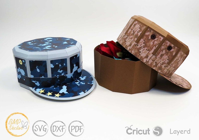 May include: Two papercraft boxes shaped like military hats. The box on the left is blue with a camouflage pattern and stars. The box on the right is brown with a camouflage pattern. Both boxes are open and have a brown interior.