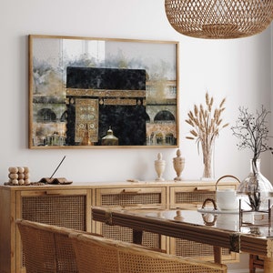 Pode incluir: Uma pintura em aquarela da Caaba em Meca, Arábia Saudita. A pintura está emoldurada em uma moldura de madeira e está pendurada em uma parede branca. A pintura é cercada por uma variedade de itens decorativos, incluindo um vaso de flores secas, uma mesa de madeira e uma cadeira de vime.