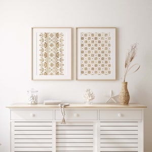 May include: Two framed prints with gold geometric patterns on a white background. The prints are hanging on a white wall above a white dresser with two drawers and two cabinet doors.