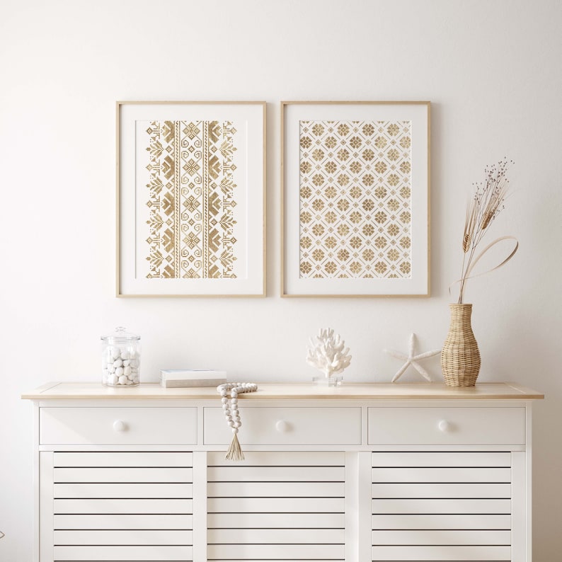 May include: Two framed prints with gold geometric patterns on a white background. The prints are hanging on a white wall above a white dresser with two drawers and two cabinet doors.