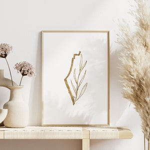 May include: A gold outline of the country of Israel with a gold leaf branch inside, framed in a light wood frame, on a light wood shelf with two vases of dried flowers and a tall vase of dried pampas grass.