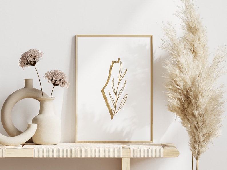 May include: A gold outline of the country of Israel with a gold leaf branch inside, framed in a light wood frame, on a light wood shelf with two vases of dried flowers and a tall vase of dried pampas grass.