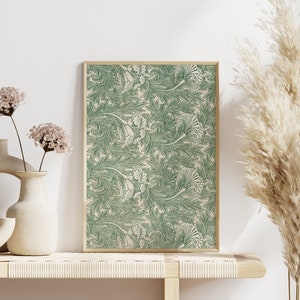 Op de afbeelding: Een ingelijst print met een groen en wit bloemenpatroon. De print hangt aan een witte muur boven een houten plank met twee vazen en een droogbloemenarrangement.