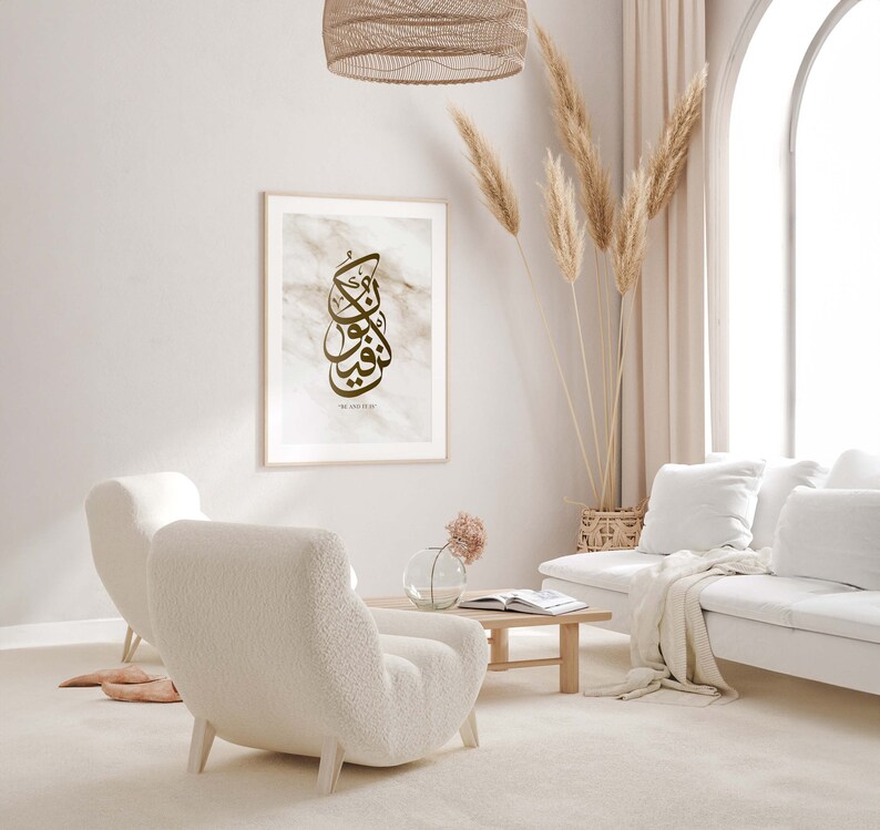 May include: A living room interior with a white couch, two white fluffy armchairs, a coffee table, a vase with pink flowers, and a framed print with Arabic calligraphy and the text "Be and Stay".