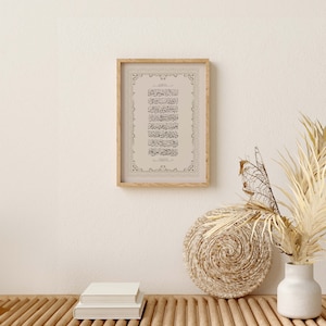 May include: A framed print of Islamic calligraphy in black ink on a beige background. The frame is made of wood and has a decorative border. The print is hanging on a white wall above a wooden bench with two books stacked on top of each other. A woven basket and a white vase with dried flowers are on the bench.