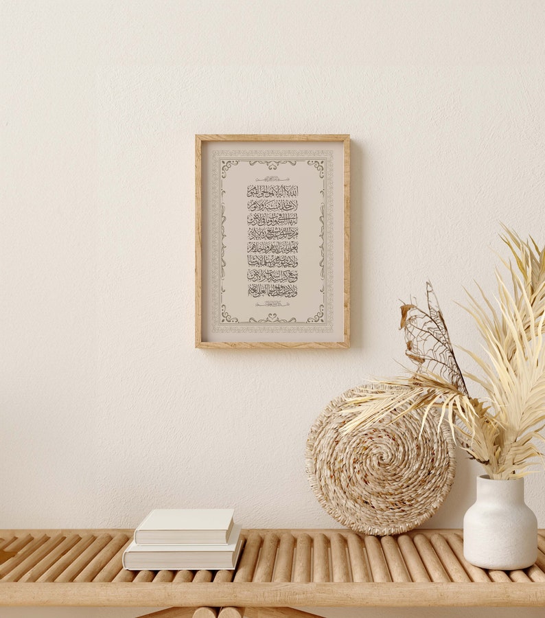 May include: A framed print of Islamic calligraphy in black ink on a beige background. The frame is made of wood and has a decorative border. The print is hanging on a white wall above a wooden bench with two books stacked on top of each other. A woven basket and a white vase with dried flowers are on the bench.