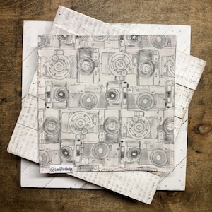 May include: A square, light-colored fabric with a repeating pattern of vintage cameras in shades of gray. The fabric is layered on top of a white, distressed wooden surface. The lower left corner of the fabric has a small tag that reads "Liberty Hanks."