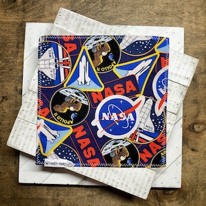 May include: A square, navy blue fabric item with a repeating pattern of NASA logos, space shuttle illustrations, and "Apollo 11" patches. The item is bordered with a decorative stitch and features the text "LIBERTY HANKS".