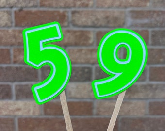 May include: Two neon green number 59 cake toppers with light blue outlines, attached to wooden sticks. The numbers are set against a brick wall background.