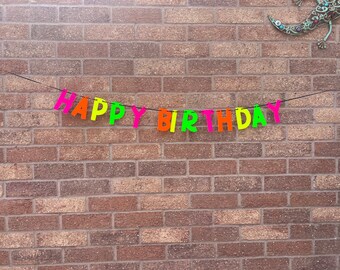 May include: A colorful "HAPPY BIRTHDAY" banner with neon pink, orange, green, and yellow letters hangs against a brick wall. The banner is strung on a thin black string, creating a festive decoration.