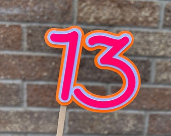 May include: A colorful number 13 cake topper. The number is hot pink with a light blue outline, set against an orange border. It is attached to a wooden stick, perfect for a birthday cake.