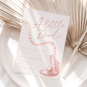 Puede incluir: Una tarjeta de invitación rectangular para baby shower con el texto A little Pearl IS ON THE WAY, Fiona and David, y detalles del evento. El diseño incluye un collar de perlas rosas sobre la tarjeta, que termina en una concha marina rosa abierta.