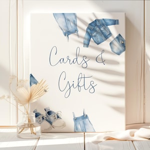 May include: A white sign with the words "Cards & Gifts" in blue script. The sign is decorated with watercolor illustrations of denim clothing items, including a jacket, overalls, and shoes. A small vase with dried flowers is in the foreground.