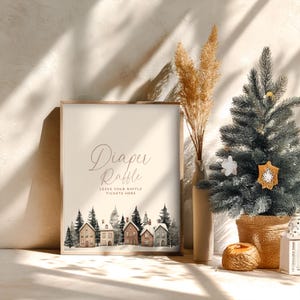 May include: A framed sign with the words "Diaper Raffle" in elegant script. Below, it reads "Leave Your Raffle Tickets Here." The bottom features a watercolor illustration of houses and trees. A small Christmas tree and vase are in the background.